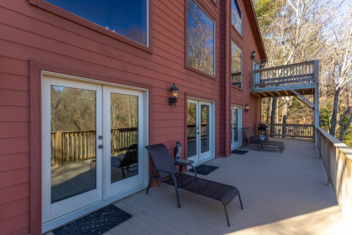 A wide exterior view of the cabin's deck area, featuring modern deck furniture and a balcony with expansive mountain views through large windows. The scene highlights opportunities for relaxing, entertaining, and connecting with the mountain landscape.