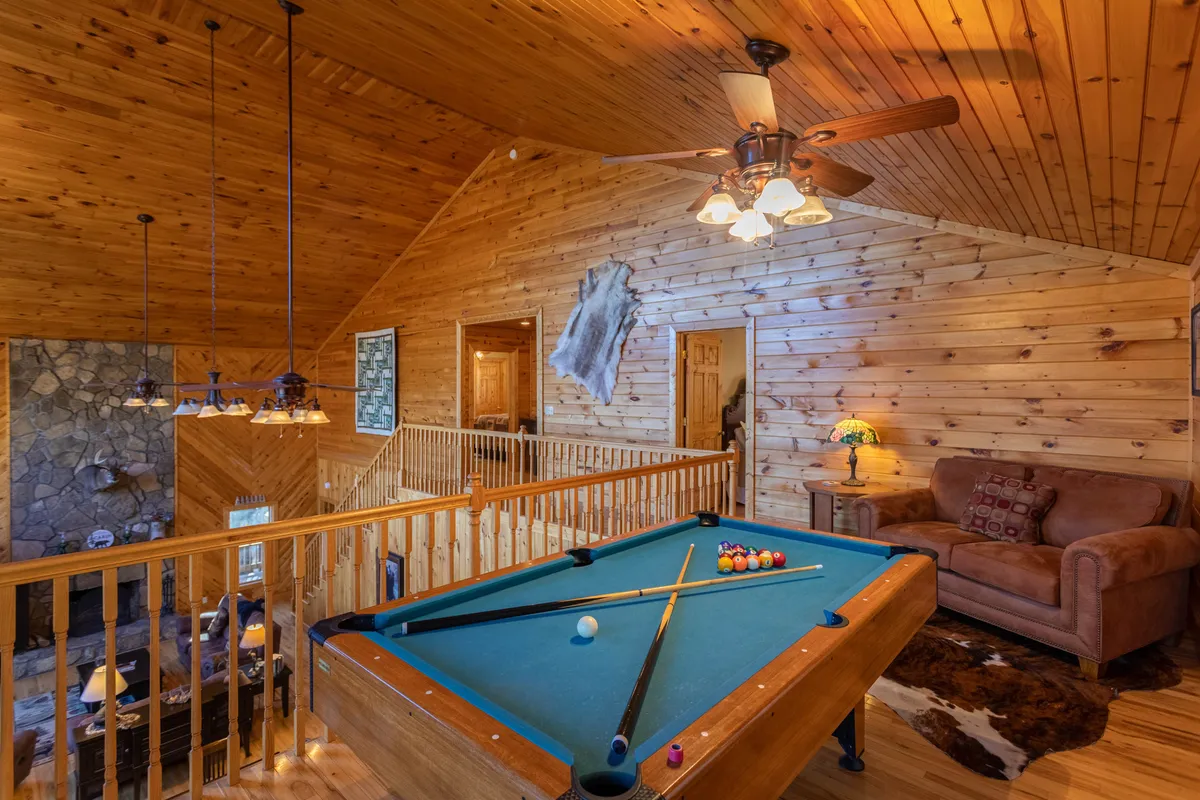 This image captures the open loft space featuring a classic pool table, a wooden railing overlooking the lower level, a stone fireplace, and a leather sofa. The setting is illuminated by natural light, creating an inviting atmosphere for socializing and friendly competition.