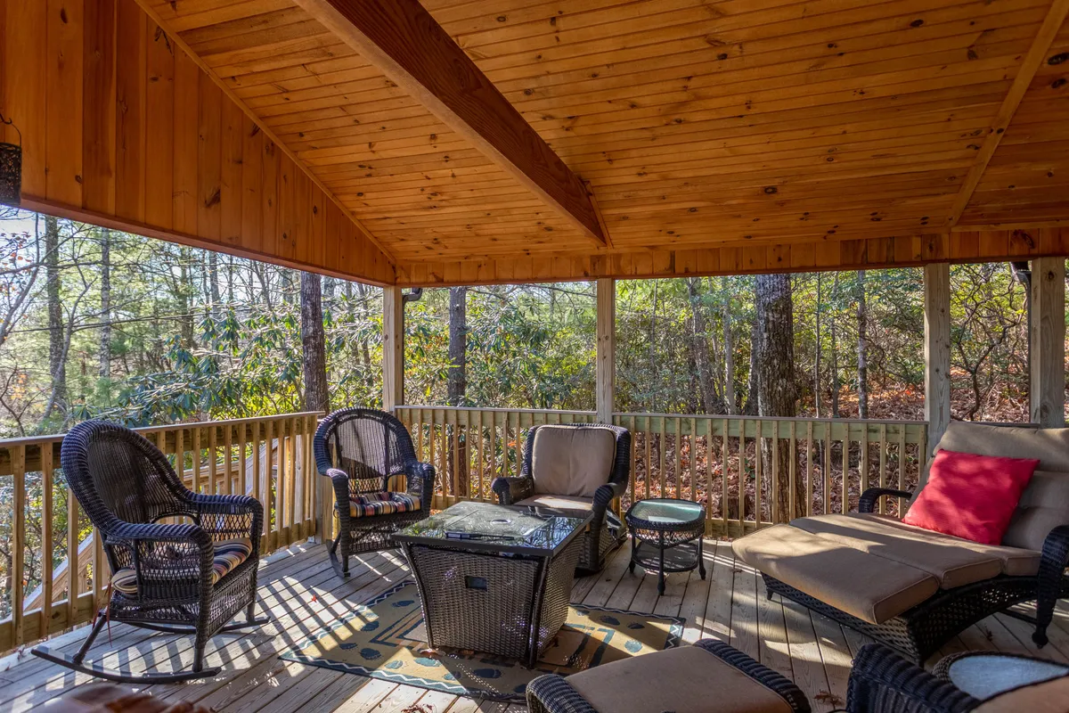 An exterior shot showing a covered deck with comfortable seating, a coffee table, and views of trees all around. The tranquil space encourages relaxed socializing, whether it's sipping cocktails at sunset, enjoying morning coffee, or cozy conversations around the table.