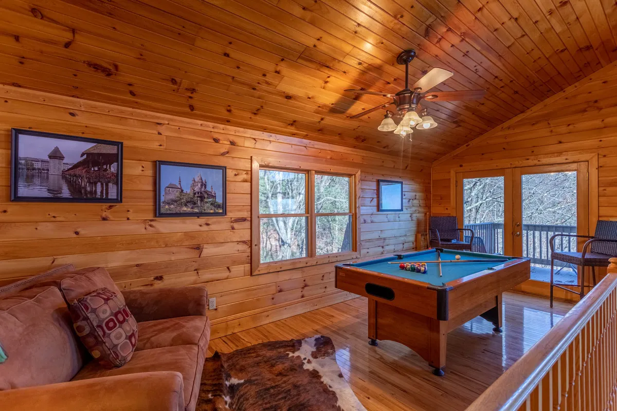 This image highlights a pool table set in a bright loft area, complemented by a comfortable sofa, windows with scenic views, and a ceiling fan above. Framed photographs on the walls add a homely touch, while the spacious layout encourages both play and relaxation.