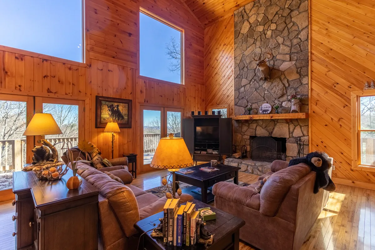 A cozy living area with a substantial stone fireplace, large windows streaming natural sunlight, plush sofas, and wooden furniture. The arrangement encourages intimate gatherings, fireside relaxation, and comfortable lounging for groups or quiet moments.
