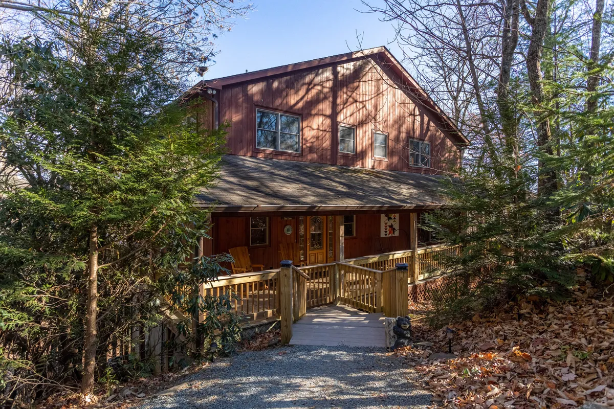 A welcoming view of the cabin's wooden deck and entrance, surrounded by lush trees. The sturdy, wrap-around deck is visible, providing a safe, inviting space for families to gather and relax after exploring the area. The natural landscape hints at the peaceful mountain setting, perfect for kids and pets to play safely.