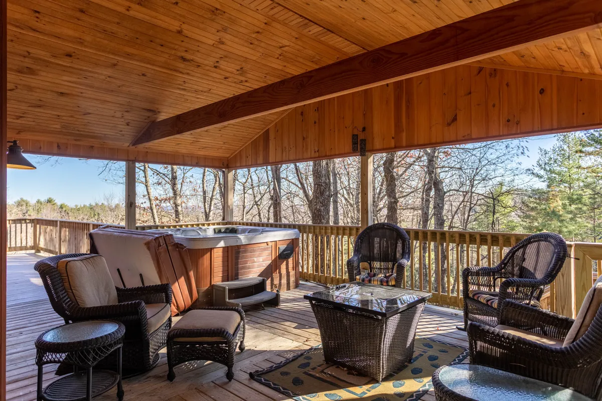 This exterior image features a spacious wooden deck with a hot tub, wicker seating, and a glass-topped table, all surrounded by nature. The area exudes relaxation and offers an inviting spot for socializing, soaking in the hot tub, or enjoying evening cocktails with friends against a serene natural backdrop.