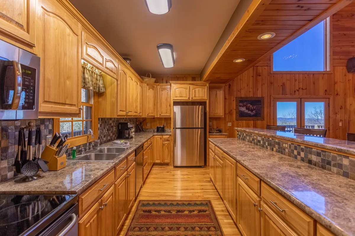 A bright, communal kitchen with wood cabinetry, granite countertops, stainless steel appliances, and large windows with scenic views. The layout includes a breakfast bar area with comfortable seating, creating an inviting atmosphere for cooking and casual dining.