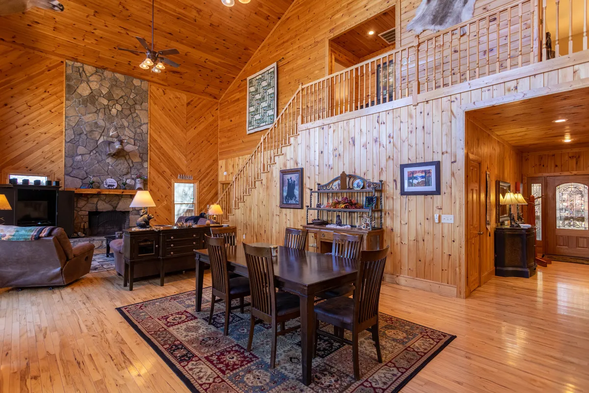 A spacious great room featuring a two-story stone wood-burning fireplace as the central focus. The room includes a large wooden dining table, a comfortable leather sectional, a wooden staircase, and expansive windows letting in natural light. The space is arranged for both relaxation and group gatherings, blending the living and dining areas seamlessly.