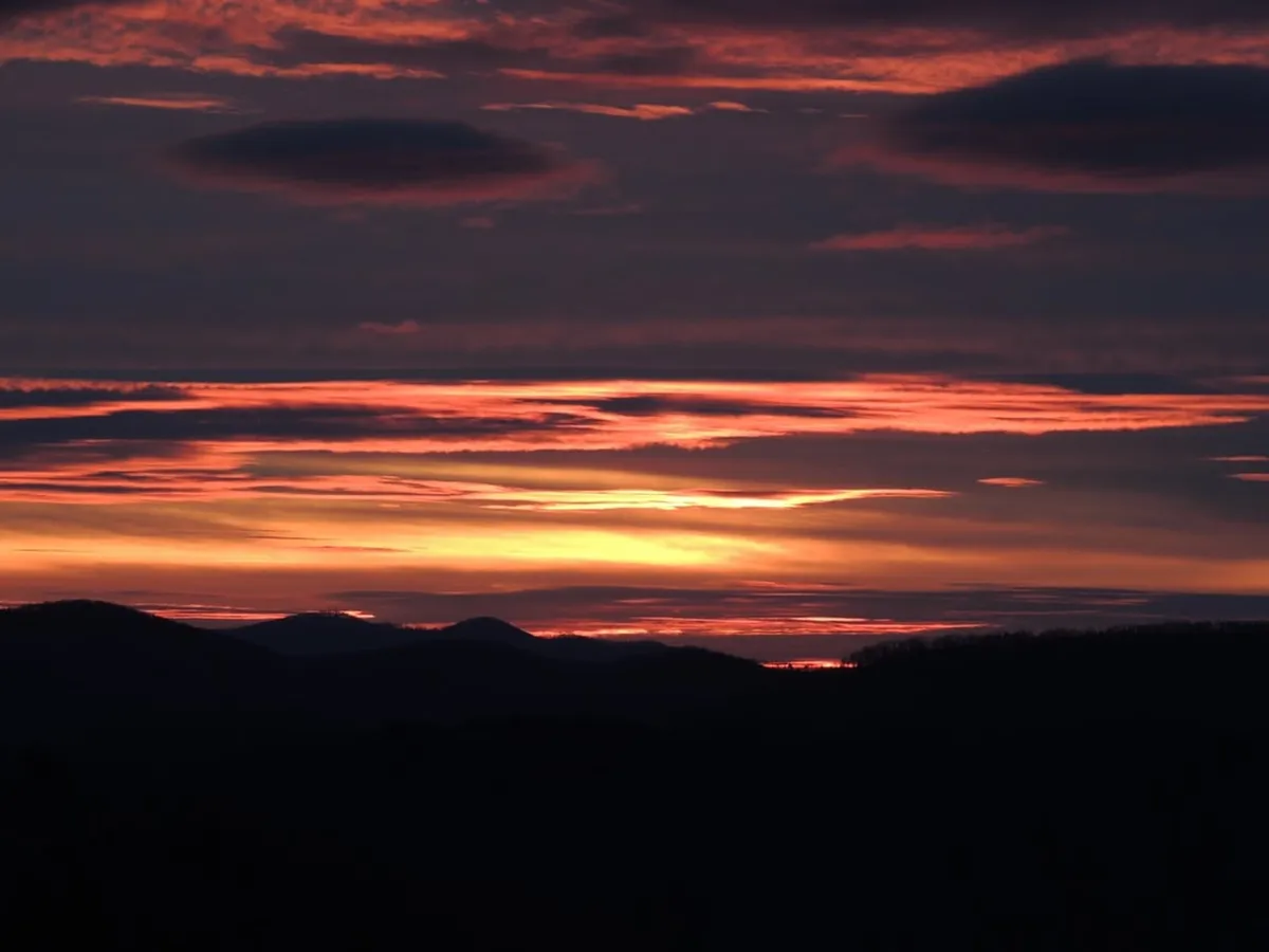 A breathtaking mountain sunset, with warm colors illuminating the sky above distant peaks. Soft clouds catch the evening light, creating a tranquil and awe-inspiring scene that captures the magic of the Blue Ridge at day's end.