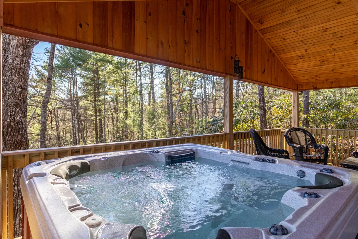 A peaceful exterior deck scene with a hot tub nestled among surrounding trees. The wooden deck is spacious, featuring seating and natural views, evoking the cozy, social side of relaxing outdoors after a day of adventure.